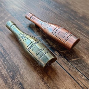 Könnte beinhalten: Zwei Miniatur-Flaschenverschlüsse aus Metall in Form von Champagnerflaschen. Die Flaschenverschlüsse haben die Form einer traditionellen Champagnerflasche mit einem Korken. Ein Flaschenverschluss ist golden und der andere ist kupferfarben. Der goldene Flaschenverschluss trägt den Text "Remy Martin" und "3CL" auf dem Etikett. Der kupferfarbene Flaschenverschluss trägt den Text "Remy Martin" und "3CL" auf dem Etikett.
