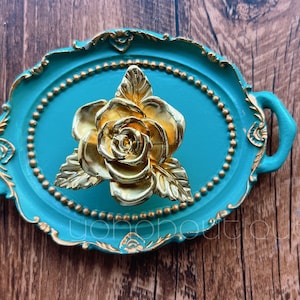 May include: A teal blue oval tray with a gold-colored rose design. The tray has a decorative gold trim and a beaded border.