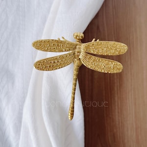 Puede incluir: Un recogedor de cortinas de metal dorado con forma de libélula. La libélula tiene alas y cuerpo detallados, y sujeta una cortina blanca. El recogedor es un elemento decorativo para la decoración del hogar.