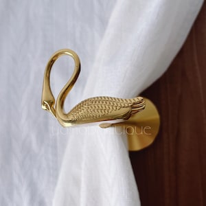 Pode incluir: Apertura de cortina dourada em forma de cisne. O cisne tem um pescoço curvo e um design detalhado de penas. A abertura é fixada a uma base redonda, segurando uma cortina branca. O fundo é de madeira.