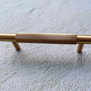 Op de afbeelding: Een gouden kastgreep met een geribbeld ontwerp. De greep heeft een eenvoudige, moderne stijl.