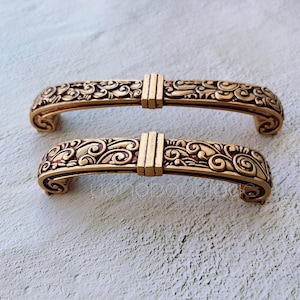 Op de afbeelding: Twee sierlijke, goudkleurige ladegrepen met ingewikkelde bloemmotieven. De grepen zijn boogvormig en hebben een decoratief middenstuk.