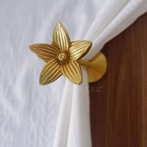 May include: A decorative gold-coloured curtain tieback in the shape of a flower. The metal tieback has five detailed petals and is holding a white curtain. The word "yonoboutique" is printed on the curtain.