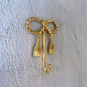 May include: A gold-coloured wall hook featuring a bow-shaped top and two tassel-like ornaments. The hook has a curved, J-shaped design. The hook is mounted on a textured, light grey wall.