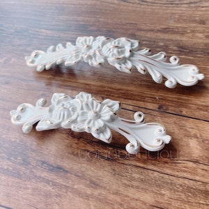 Peut inclure: Deux poignées de tiroir en céramique blanche avec des accents dorés. Les poignées sont en forme de fleurs stylisées avec des détails complexes.