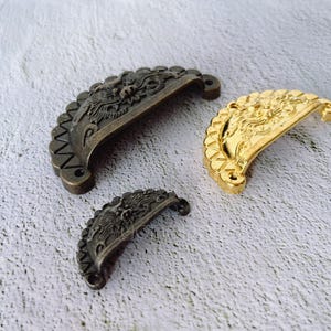 Könnte beinhalten: Drei dekorative Schubladengriffe in Bronze und Gold. Die Griffe haben eine Halbmondform mit verzierten Drachendesigns. Die Bronzegriffe haben eine dunkle, antike Oberfläche, während der goldene Griff glänzend ist. Für Möbel.