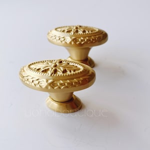 Pode incluir: Dois botões de armário ovais dourados com um design floral ornamentado. Os botões têm um centro elevado e um acabamento liso e polido.