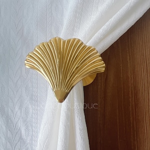 Puede incluir: Un recogedor de cortina dorado en forma de abanico. El recogedor sujeta una cortina blanca. El fondo incluye una puerta de madera y una pared blanca texturizada. El recogedor tiene un diseño decorativo y acanalado.