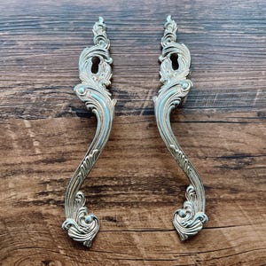 May include: Two ornate silver door handles with a keyhole in the center. The handles have a decorative, scroll-like design.