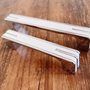 May include: Two silver cabinet door pulls with a rectangular shape and a slightly raised, rectangular design in the center.