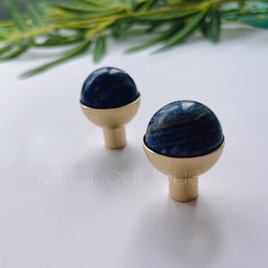 Könnte beinhalten: Zwei goldene Schrankknöpfe mit blauen Lapislazuli-Steinoberflächen. Die Knöpfe sind rund und haben eine glatte, polierte Oberfläche.