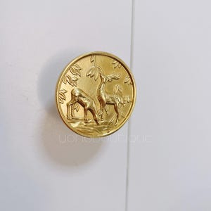 Op de afbeelding: Een ronde kastknop in goudkleur met een reliëf van twee herten in een boslandschap.