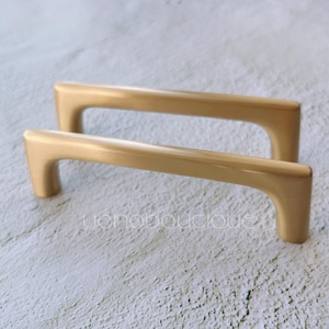 Könnte beinhalten: Zwei goldene Schrankgriffe mit einem schlichten, modernen Design. Die Griffe sind aus Metall und haben eine glatte, polierte Oberfläche.