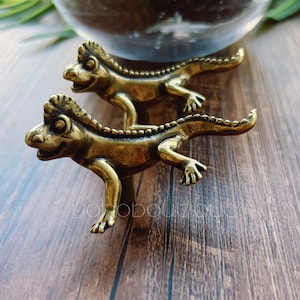 Peut inclure: Deux figurines de lézard en laiton antique. Chaque lézard a un corps détaillé et texturé avec un visage souriant et de petites pattes. Les lézards sont placés sur une surface en bois, suggérant un objet décoratif ou de collection.