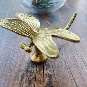 Puede incluir: Un pomo o tirador de puerta en forma de libélula de color dorado. La libélula tiene alas detalladas y una cola larga.