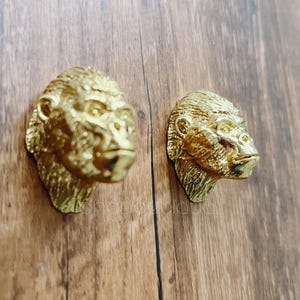 May include: Two gold-coloured gorilla head door knobs. The gorilla heads are facing to the right. The knobs are mounted on a wooden surface.