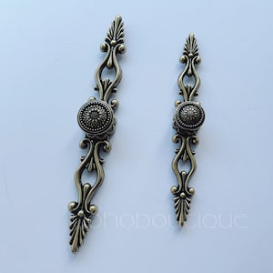 Może przedstawiać: Dwa ozdobne brązowe uchwyty do drzwi szafki z kwiatowym wzorem. Każdy uchwyt ma okrągły gałkę z kwiatowym wzorem.