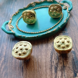 May include: Four gold-toned lotus flower shaped cabinet knobs with a raised design. The knobs are displayed on a teal tray with a gold rim.