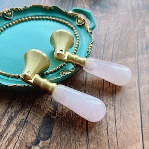 Pode incluir: Dois botões de armário dourados com alças cor-de-rosa em forma de lágrima. Os botões são exibidos em uma bandeja turquesa com detalhes dourados.