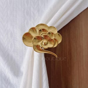 Può includere: Un fermatenda dorato a forma di nuvola. Tiene una tenda bianca. Il design presenta un centro a spirale e molteplici forme di nuvola. Il testo "yomoboutique" è visibile.