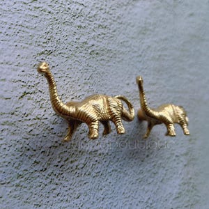 Puede incluir: Dos figuritas de dinosaurio de color dorado, una de pie y la otra tumbada, están montadas en una pared blanca.