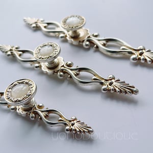 Puede incluir: Tres pomos de armario plateados con diseños ornamentados y centros de color blanco perlado.