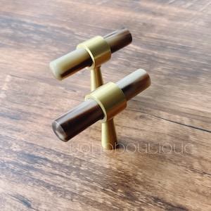 Op de afbeelding: Twee gouden kastknoppen met bruine en witte marmeren accenten. De knoppen worden getoond op een houten oppervlak.