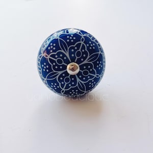 以下が含まれることがあります： 白い花の模様が施された青いセラミック製のノブ。ノブの中央には銀色の金属製のアクセントがあります。