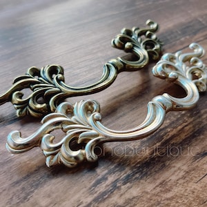 Op de afbeelding: Twee sierlijke, goudkleurige ladegrepen met een decoratief bloemmotief. De grepen hebben de vorm van een halve cirkel met een puntig uiteinde.