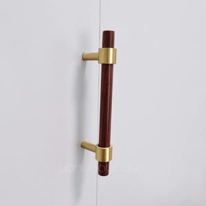 Może przedstawiać: Drewniany uchwyt do szafki w kolorze ciemnobrązowym z dwoma złotymi metalowymi pierścieniami na końcach. Uchwyt jest zamontowany na białej powierzchni.