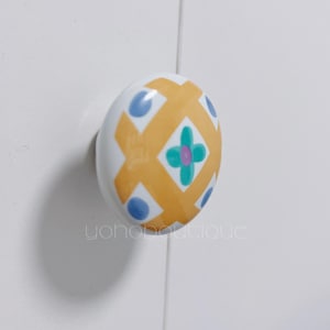 Könnte beinhalten: Ein weißer Keramik-Schrankknopf mit einem farbenfrohen geometrischen Design. Das Design zeigt ein gelbes Kreuzschraffurmuster, blaue Punkte und ein türkis-violettes Blumenmuster. Der Text "yohoboutique" ist sichtbar.