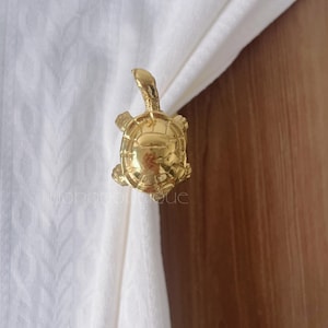 Könnte beinhalten: Ein goldfarbener, schildkrötenförmiger Vorhanghalter. Die Schildkröte hat einen detaillierten Panzer, Kopf und Flossen. Der Vorhanghalter ist an einem weißen Vorhang mit Holzgrund befestigt.