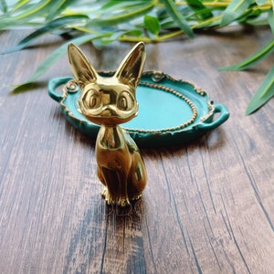 以下が含まれることがあります： 金色のかわいらしい猫の置物が、ターコイズブルーの縁取りが施された金色のトレーの上に置かれています。トレーには2つの取っ手が付いており、木製の表面の上に置かれています。