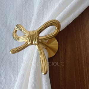 Peut inclure: Un embrasse-rideau en métal doré en forme de nœud. L'embrasse-rideau maintient un rideau blanc. L'arrière-plan est une surface en bois.