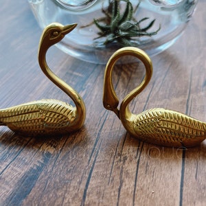 May include: Two brass swan ornaments, one with its head raised and the other with its head curved down. The swans have detailed feather patterns and are on a wooden surface. A glass vase with greenery is in the background.