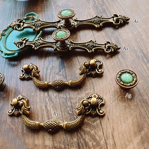 May include: Antique brass cabinet hardware with green stone accents. The collection includes ornate pulls and knobs with detailed floral and lion head designs. The hardware is displayed on a wooden surface.