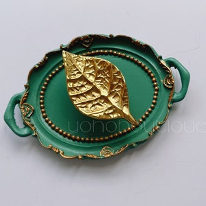 May include: A green oval tray with gold accents and a gold leaf design in the center. The tray has two handles on the sides.