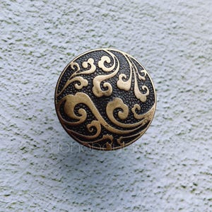 May include: A circular button with an antique aesthetic. The button has a dark, textured base with raised, swirling patterns in a gold hue. Likely made of metal, it's suitable for sewing or craft projects. The design is reminiscent of vintage styles.