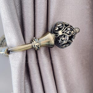 Może przedstawiać: Złoty metalowy zaczep do zasłon z ozdobnym wzorem. Zaczep trzyma parę zasłon.