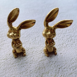 以下が含まれることがあります： 長い耳を持つ金色のウサギの置物2つ。ウサギは直立して、前足を交差させて立っています。