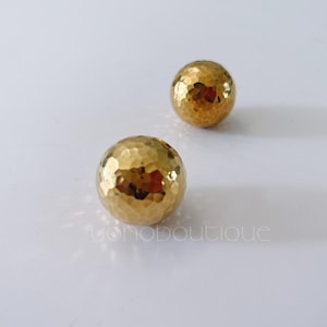 Op de afbeelding: Twee gouden metalen bollen met een gehamerde textuur. De bollen liggen op een wit oppervlak.