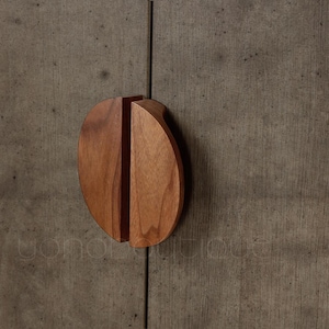 Pode incluir: Puxador de armário de madeira com acabamento castanho-avermelhado quente. O puxador tem formato oval arredondado com uma fenda vertical no centro. O fundo é uma superfície cinza texturizada.