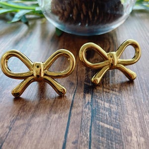 May include: Two gold-colored bow-shaped decorative objects. The bows are symmetrical with rounded loops and a knot in the center. They are placed on a dark wooden surface, with a glass container and greenery in the background.