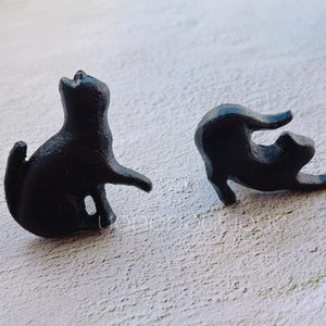 Puede incluir: Dos figuritas de gato negro, una de pie y la otra tumbada de lado, ambas con un acabado liso y elegante.