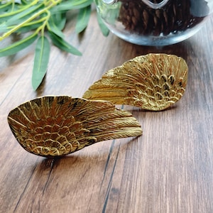 Könnte beinhalten: Zwei goldfarbene, flügelförmige Dekorationsgegenstände. Die Flügel sind mit federartigen Texturen versehen und befinden sich auf einer Holzoberfläche. Die Flügel werden wahrscheinlich zu Dekorationszwecken verwendet.