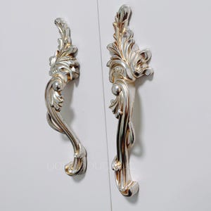 May include: Two ornate silver cabinet door pulls with a floral design. The pulls are attached to a white surface.