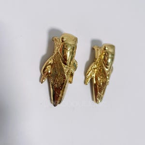 Pode incluir: Dois brincos dourados em forma de espiga de milho. Os brincos têm uma textura detalhada e estão colocados sobre um fundo branco. São provavelmente feitos de metal e projetados como joias.