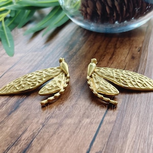 May include: A pair of gold-coloured dragonfly-shaped earrings. The earrings have detailed wings and a segmented body. The wings have a textured pattern. The earrings are displayed on a wooden surface.