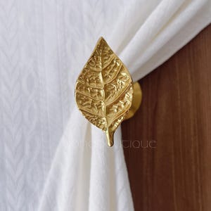 Peut inclure: Un embrasse-rideau doré en forme de feuille. L'embrasse est en métal et présente des nervures détaillées. La feuille maintient un rideau blanc. L'arrière-plan est une porte en bois.