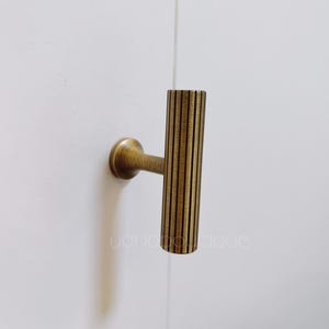 Könnte beinhalten: Ein goldfarbener Metall-Schranktürgriff mit vertikalem Rippenmuster. Der Griff ist an einer weißen Wand montiert.